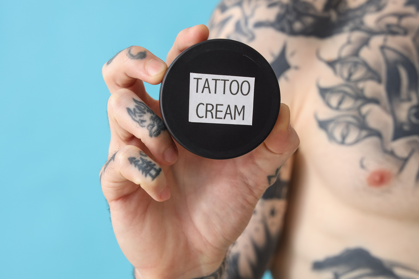 Tattooed Man Holding Jar of Tattoo Cream on Blue Background, Closeup